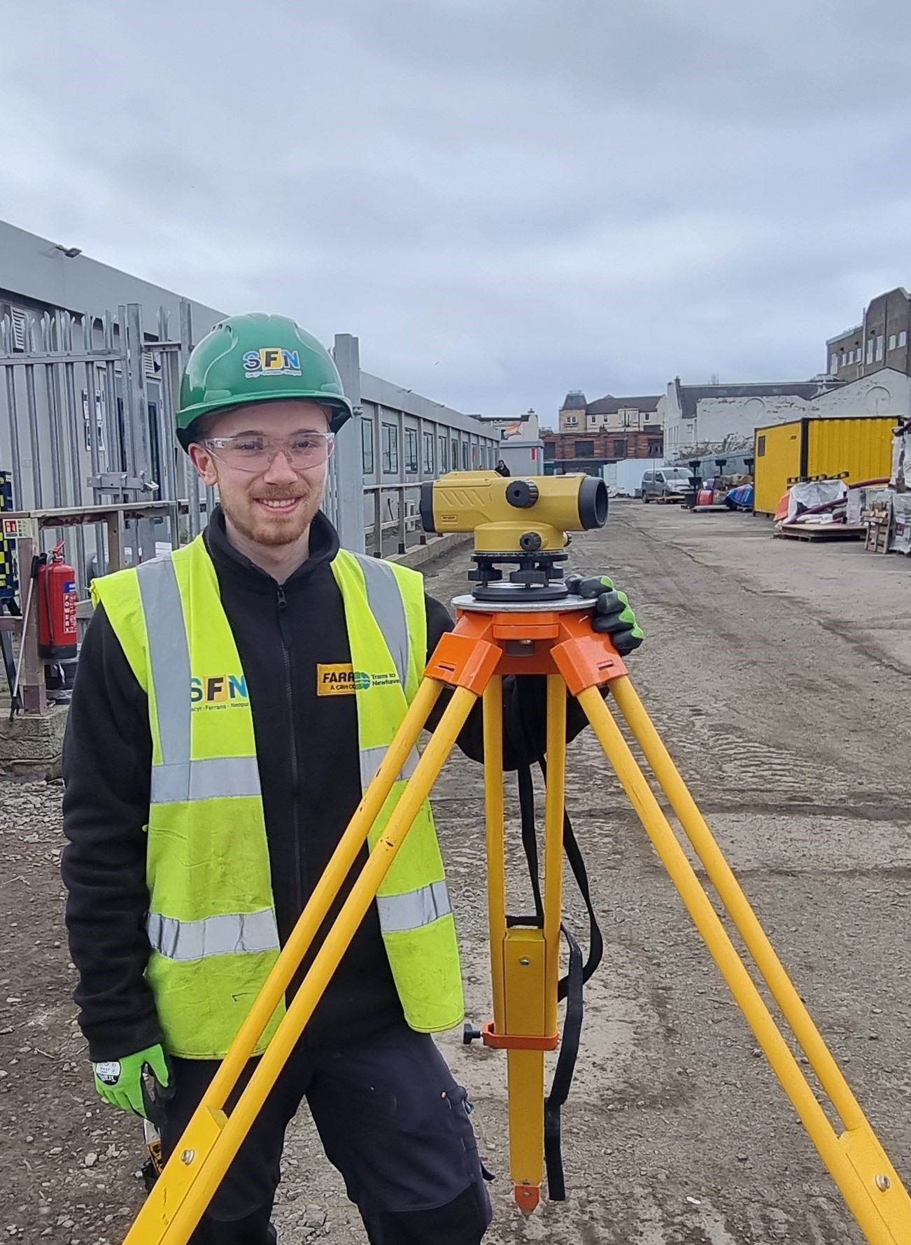 Photo of smiling apprentice in yellow high viz jacket posing on site next to equipment.