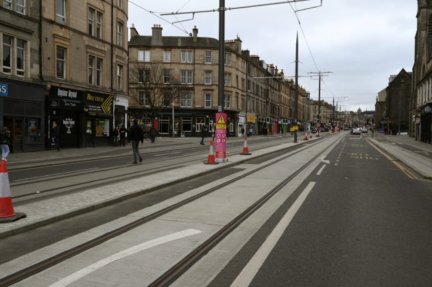 Photo of Leith Walk just before its junction with Iona Street. The OLE poles bearing the energised cables can be seen in the foreground.